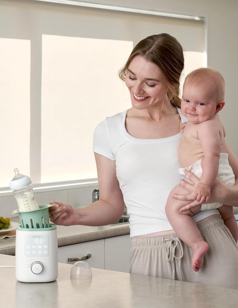 A mother stands in a kitchen, holding her baby close with one arm while using her free hand to operate a bottle warmer for feeding.