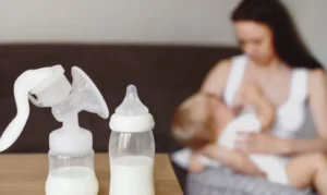A manual breast pump and a baby bottle filled with milk sit on a table, illustrating combination feeding equipment. In the blurred background, a mother is nursing her baby directly, highlighting the mix of breastfeeding and bottle-feeding.