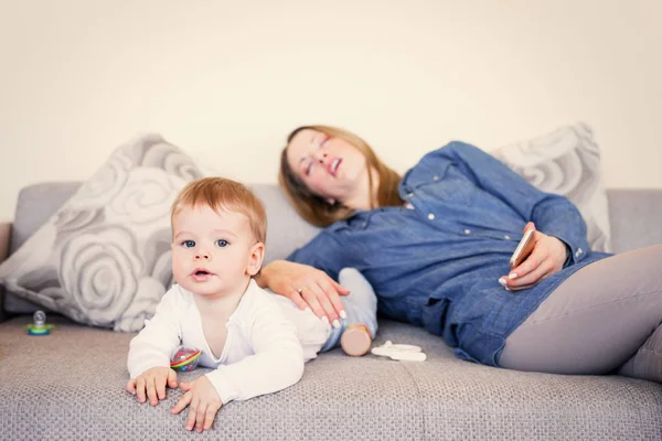 An exhausted mother sleeping on a couch while holding a smartphone, with her wide-awake, alert baby lying next to her, capturing the exhaustion of the 4-month sleep regression.