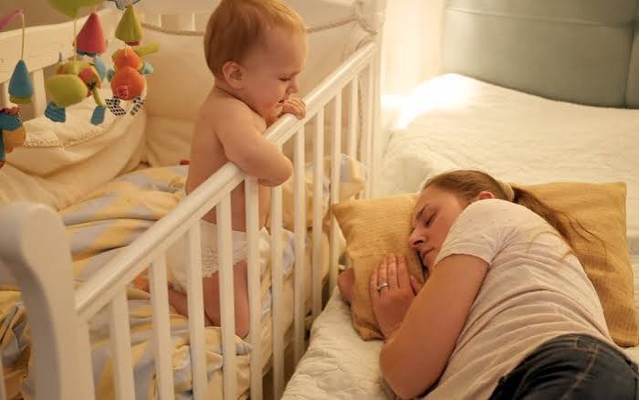 An exhausted mother sleeping next to a crib while an awake older baby stands up, illustrating the physical toll of a newborn waking every hour at night.