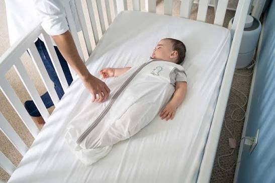 A parent zipping an infant into a sleep sack inside a crib, illustrating how to transition from swaddle to sleep sack safely.